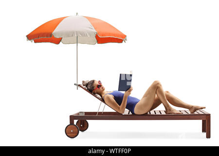 Full length profile shot of a young woman in a swimsuit lying on a sunbed with umbrella reading a book isolated on white background Stock Photo