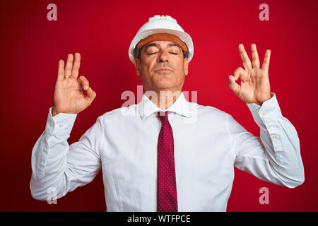 Handsome middle age engineer man wearing security helmet over isolated red background relax and smiling with eyes closed doing meditation gesture with Stock Photo