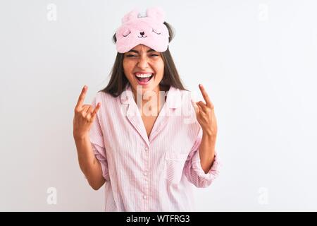 Young beautiful woman wearing sleep mask and pajama over isolated white background shouting with crazy expression doing rock symbol with hands up. Mus