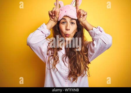 Young woman wearing pajama and sleep mask standing over yellow isolated background doing funny gesture with finger over head as bull horns