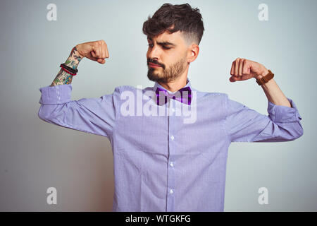 Young man with tattoo wearing purple shirt and bow tie over isolated white background showing arms muscles smiling proud. Fitness concept. Stock Photo