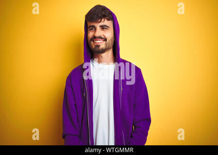 Young man with tattoo wearing purple sweatshirt with hood over isolated yellow background looking away to side with smile on face, natural expression. Stock Photo