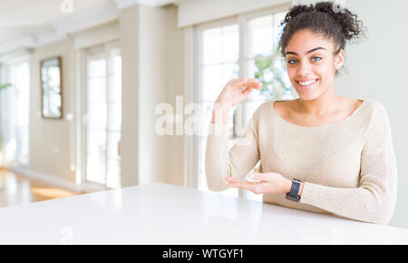 Beautiful young african american woman with afro hair sitting on table at home gesturing with hands showing big and large size sign, measure symbol. S Stock Photo