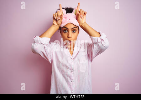 Young african american woman wearing pajama and mask over isolated pink background doing funny gesture with finger over head as bull horns
