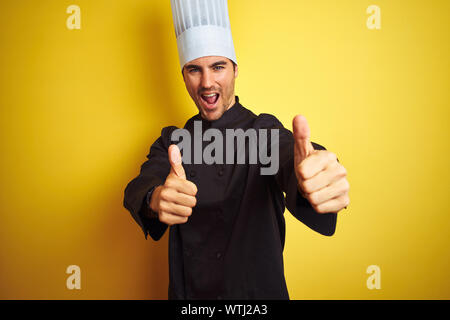 Young chef man wearing uniform and hat standing over isolated yellow background approving doing positive gesture with hand, thumbs up smiling and happ Stock Photo