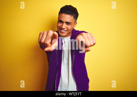 Young brazilian man wearing purple sweatshirt standing over isolated yellow background pointing to you and the camera with fingers, smiling positive a Stock Photo