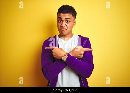 Young brazilian man wearing purple sweatshirt standing over isolated yellow background Pointing to both sides with fingers, different direction disagr Stock Photo