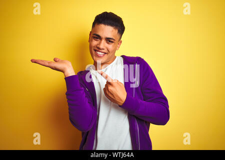 Young brazilian man wearing purple sweatshirt standing over isolated yellow background amazed and smiling to the camera while presenting with hand and Stock Photo