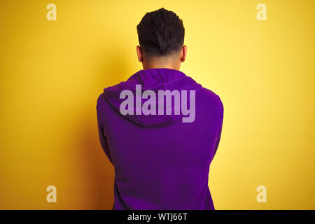 Young brazilian man wearing purple sweatshirt standing over isolated yellow background standing backwards looking away with crossed arms Stock Photo