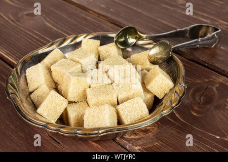 Lot of whole sweet brown sugar cube in metal bowl with sugar tongs on brown wood Stock Photo