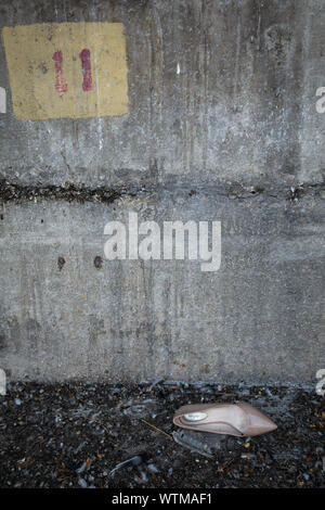 A single beige high heel shoe abandoned on the ground in front of a weathered concrete wall with a yellow sign displaying the number eleven Stock Photo