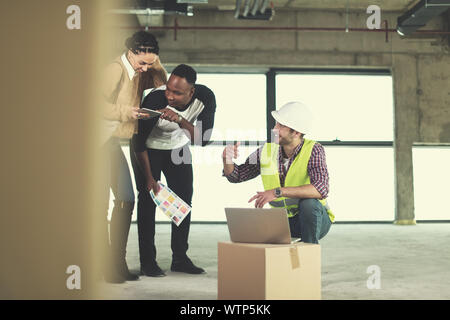 architect engineer using laptop computer while showing house design plans to a young multiethnic couple at construction site Stock Photo