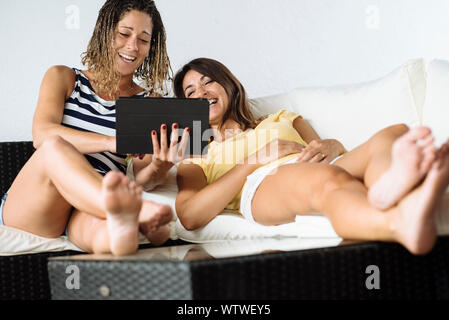 Two women laughing while looking at a tablet computer lying on a sofa. Stock Photo