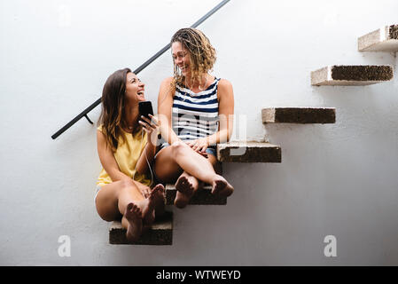 Two women sitting on stairs laughing while listening to music. Stock Photo