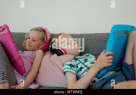siblings sat at home on the coach playing on their iPads Stock Photo