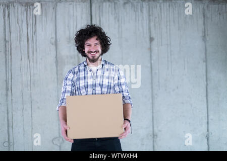 young successful smiling casual businessman carrying cardboard box in front of a concrete wall during moving in at new startup office Stock Photo