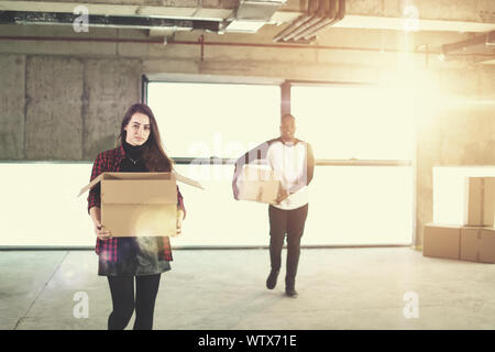 happy young multiethnic casual business team carrying cardboard boxes with sunlight through the windows during moving in at new unfinished startup off Stock Photo