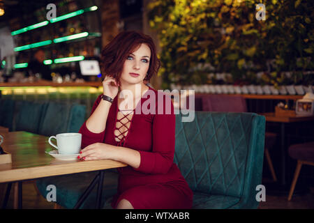 Fall casual fashion, elegant everyday look. Plus size model. Beautiful young woman in a chic red dress with a deep neckline drinking tea in a restaura Stock Photo