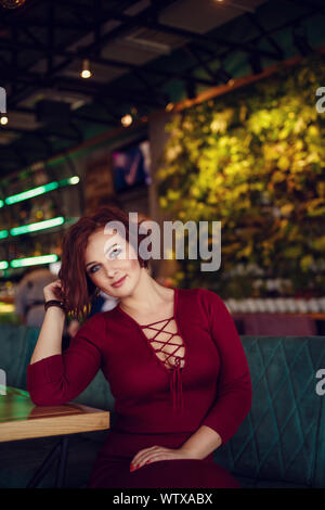 Fall casual fashion, elegant everyday look. Plus size model. Beautiful young woman in a chic red dress with a deep neckline drinking tea in a restaura Stock Photo