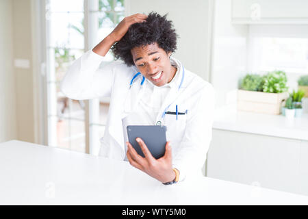 African American doctor man using tablet at the clinic stressed with hand on head, shocked with shame and surprise face, angry and frustrated. Fear an Stock Photo