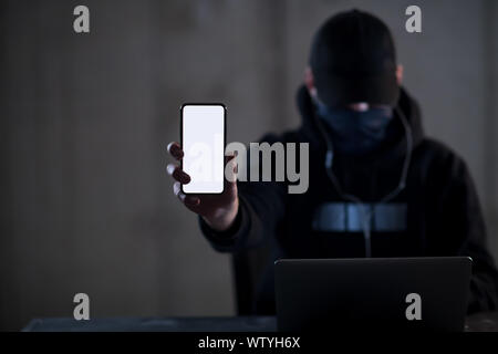 Young talented criminal hacker using laptop computer and showing the smart phone which is pirated while working in dark office with concrete wall in t Stock Photo