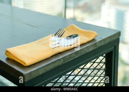 Fork, spoon and knife in an orange color napkin on a wooden table with a view of outdoor Stock Photo