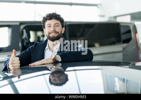 Cheerful car salesman smiling showing thumbs up posing at the dealership salon leaning on a new car for sale Stock Photo