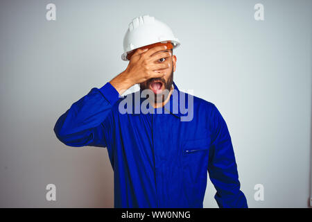 Handsome indian worker man wearing uniform and helmet over isolated white background peeking in shock covering face and eyes with hand, looking throug Stock Photo