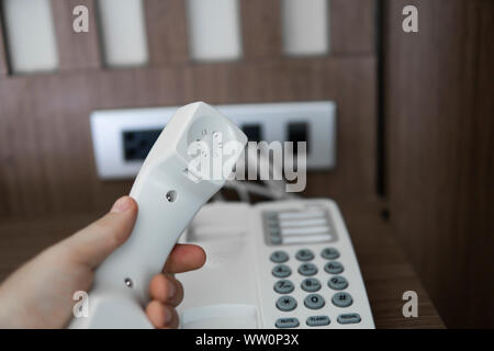 Mans hand holding handset of white vintage telephone with grey battoms on a brown table in hotel. Stock Photo