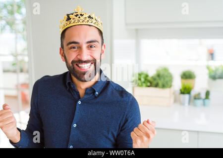 Handsome hispanic man wearing golden crown over head as the king celebrating surprised and amazed for success with arms raised and open eyes. Winner c Stock Photo