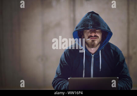 Young talented hacker smoking cigarette while working on laptop computer in dark office with concrete wall in the background Stock Photo