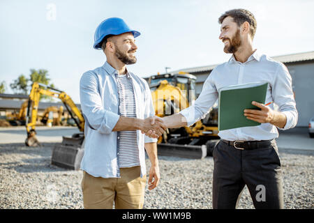 Builder choosing heavy machinery for construction with a sales consultant shaking hands on the open ground of a shop with special vehicles Stock Photo