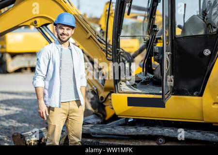 Portrait of a handsome builder standing near the escavator on the open ground of the shop with heavy machinery Stock Photo