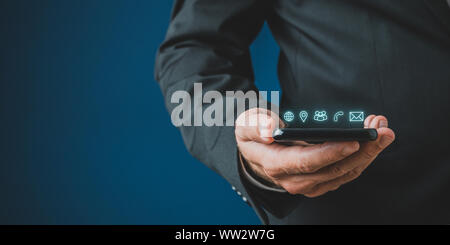 Businessman holding mobile phone with contact and communication icons glowing above it in a conceptual image. Over navy blue background. Stock Photo