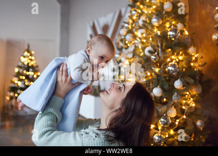 Beautiful happy mother with her little daughter in knit sweater sitting on the background of Christmas interior hugging together and smiling Stock Photo