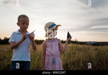 brother and sister playing with paper planes in a meadow at sunset Stock Photo