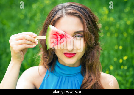 Triangle of watermelon on a stick in the hands on the girl's eye on a green background. Sweet summer weekend. Stock Photo