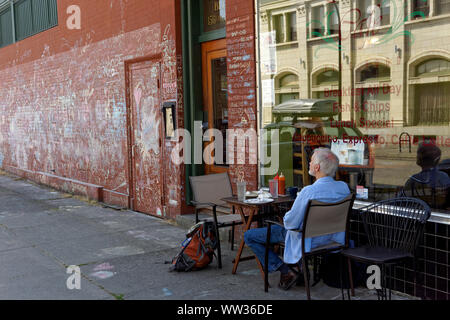 Older man sitting alone at a table outside a restaurant cafe in the Mount Pleasant district of Vancouver, BC, Canada Stock Photo