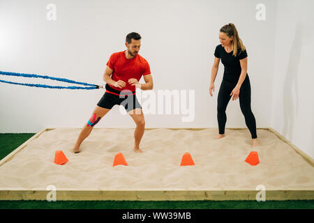 Man does stretching exercises on sand with a blue support at the waist, assisted by a physiotherapist girl, a white wall in the background. Concept of Stock Photo