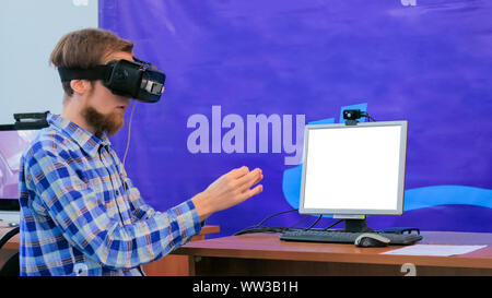 Young man using virtual reality headset in front of white blank monitor Stock Photo
