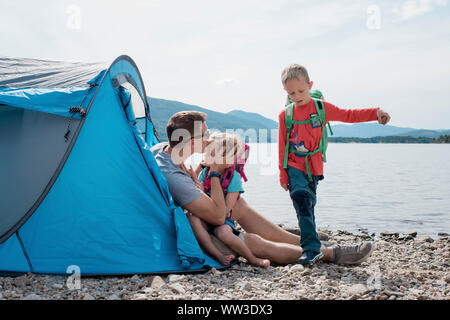 father kissing his daughter whilst his family plays camping by the sea Stock Photo