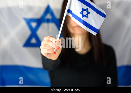 Young Woman Holding Small Israel Flag In Front Of Face on Israeli Flag Background. Copy space. Selective focus on flag . Stock Photo