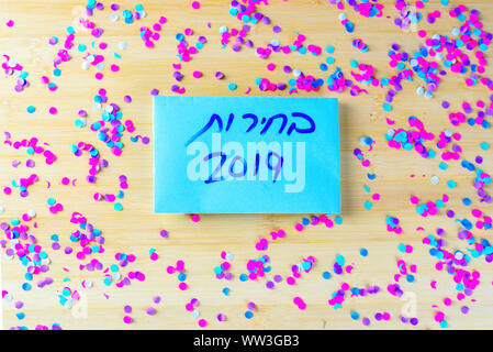 Hebrew text Elections 2019 on voting paper over wooden board with confetti background. Israel new Elections 17 September 2019. Stock Photo