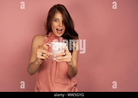 A girl in a pink top is standing on a pink background joyfully opening her mouth and holding out hands with a mug to the camera Stock Photo