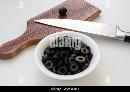 Sliced black olives in a bowl near brown wooden cutting board and chef knife on a white kitchen table. Vegetables, vegetarian food and healthy eating. Stock Photo