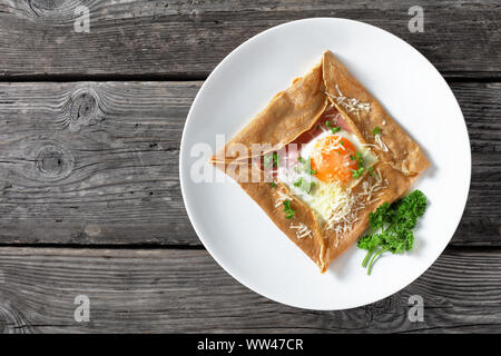 Breton crepe, Savory Buckwheat Galettes Bretonnes with fried egg, cheese, ham served on a white plate on a rustic wooden table, classic french brunch, Stock Photo