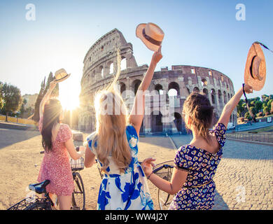 Three happy young women friends tourists with bikes waving hats at Colosseum in Rome, Italy at sunrise. Stock Photo