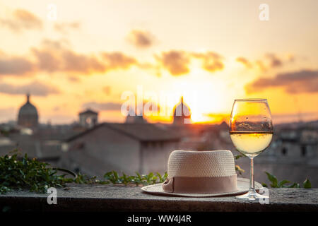 Hat, glass and bottle of white wine in front of panoramic view of Rome cityscape from campidoglio terrace at sunset. Landmarks, domes of Rome, Italy. Stock Photo