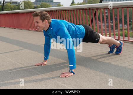 Man in blue tracksuit top doing push-ups on a bridge Stock Photo