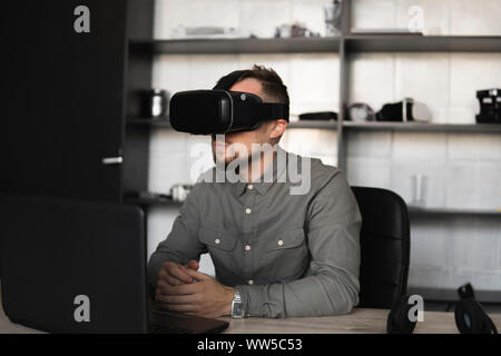 Young bearded man sitting against computer with virtual reality glasses. Modern technologies. The concept of future technology. Stock Photo